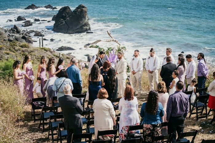 Outdoor wedding ceremony by the ocean, with guests standing, five groomsmen, and bridesmaids in lavender dresses.