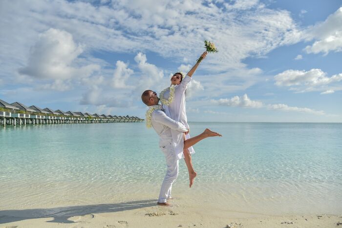 Bride and groom joyfully celebrating on a tropical beach under a bright blue sky.