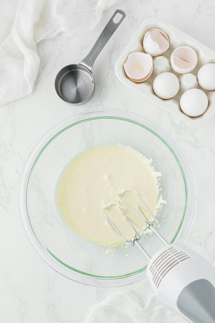 Mixer blending ingredients in a bowl, with eggshells and a measuring cup nearby.