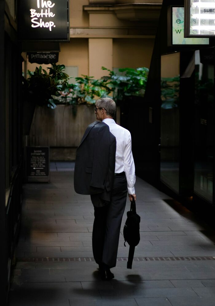 Man in a suit walking down an alley, holding a jacket over his shoulder, near a store sign, reflecting independence from parental norms.