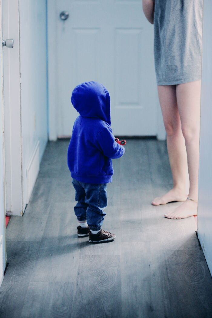 Child wearing a blue hoodie standing in a hallway, facing an adult’s legs.