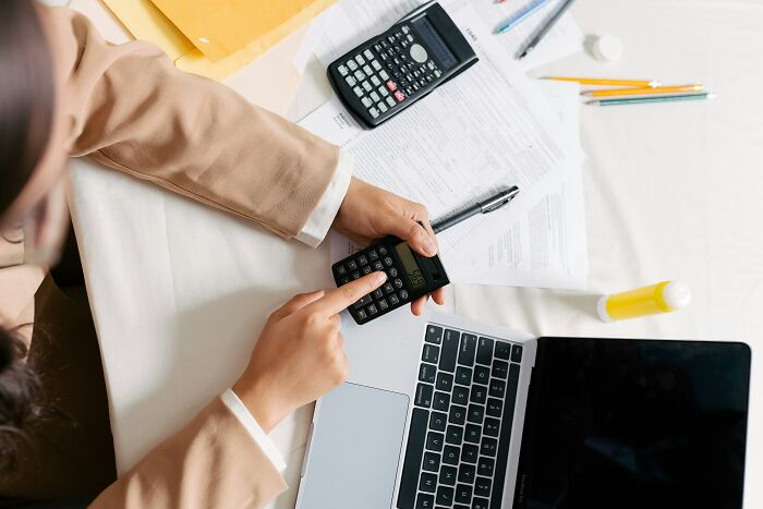 A person using a calculator with documents and a laptop on a desk, representing a task they won't repeat like their parents.