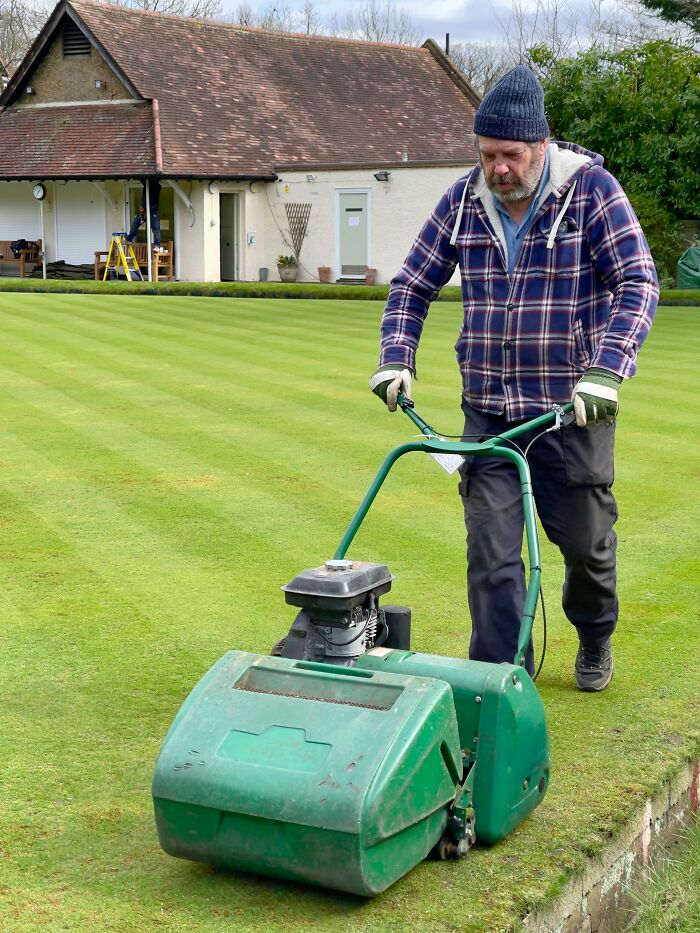 Man mowing lawn with green machine, wearing plaid jacket, beanie, and gloves, in front of a small building.