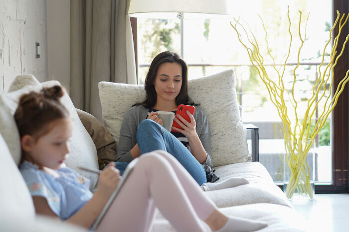 A woman holding a mug and phone, sitting on a couch, next to a child using a tablet, highlighting a relaxed setting.