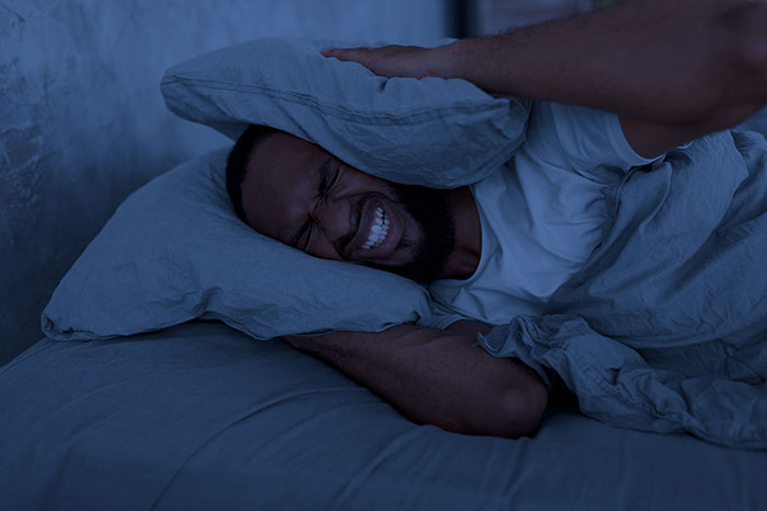 Man in bed covering ears with pillow due to hotel room neighbors' noise disturbance.