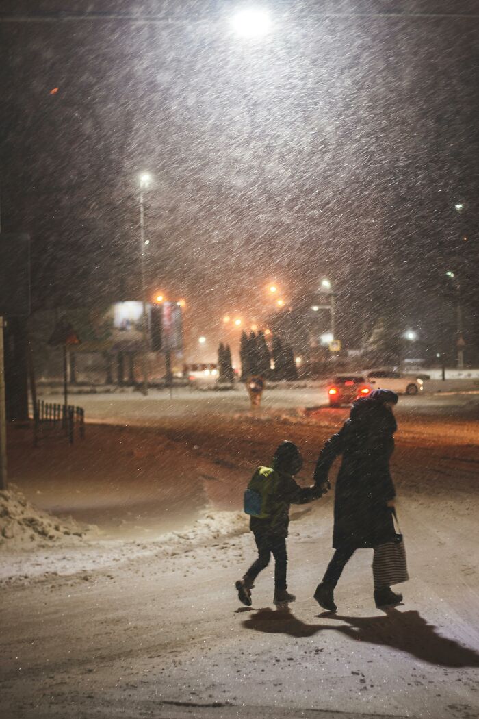 Parent holding child's hand walking in snow, illuminated by streetlights, showcasing parenting hacks in winter weather.