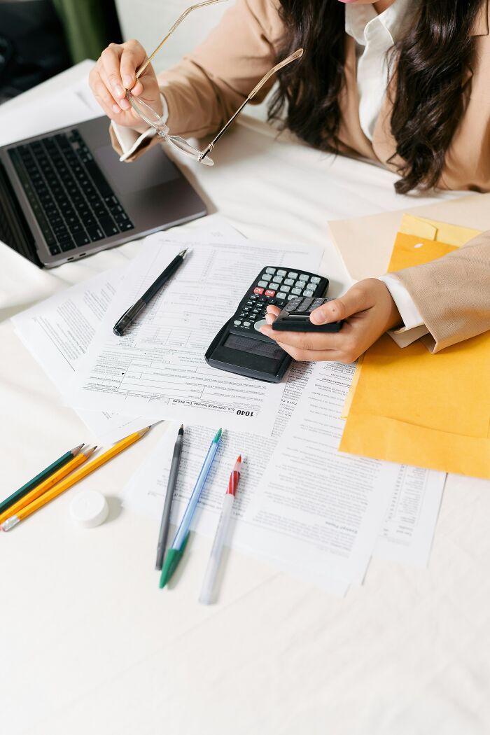 Person managing finances, symbols of adulthood challenges on table with calculator, paperwork, and laptop.