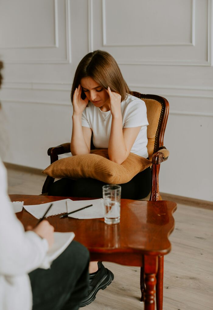Woman in a chair holding her head, representing challenges of adulthood, in a therapy session setting with papers and water.