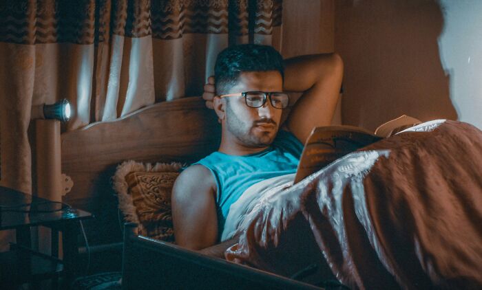 Man wearing glasses, reading in bed, surrounded by soft lighting, enhancing his life with a book, relaxation setting.