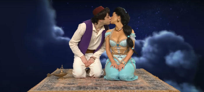 Pete Davidson and Kim Kardashian in costume kissing on a magic carpet against a night sky backdrop, related to love story rumors.