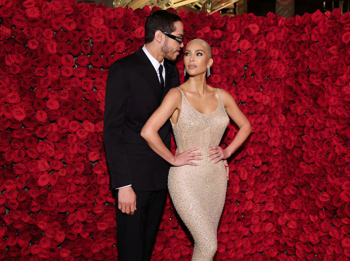 Pete Davidson and Kim Kardashian posing in front of red rose backdrop, wearing formal attire, related to their rumored love story.