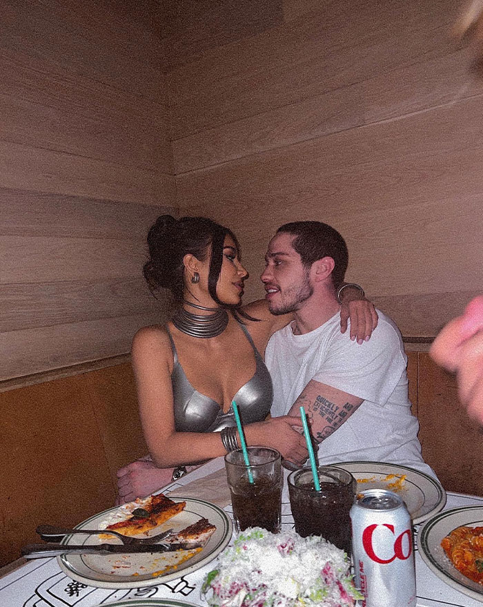 Pete Davidson and Kim Kardashian sitting closely in a cozy setting with food and drinks on the table, enjoying each other's company.