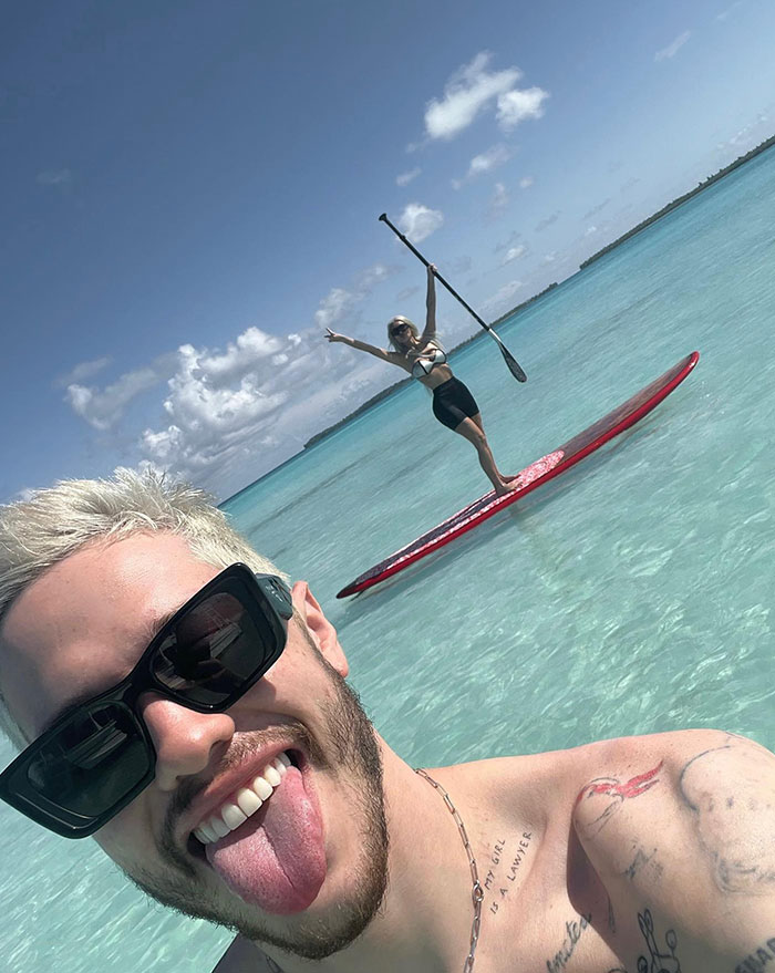 Pete Davidson smiling with sunglasses, while enjoying a sunny day at the beach with Kim Kardashian paddleboarding in the background.