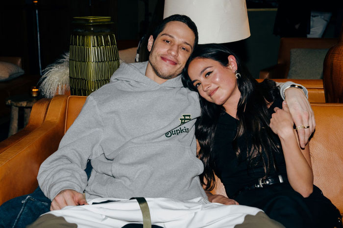 Pete Davidson and Chase Sui Wonders sitting closely on a couch, one wearing a gray hoodie, in a cozy setting.
