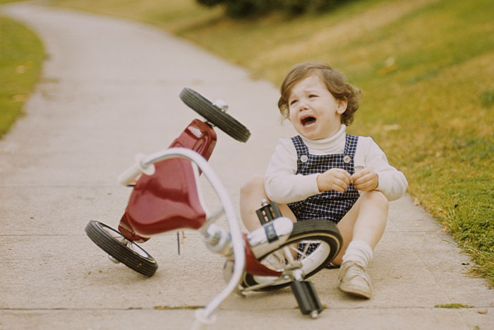 Child holding a petty grudge, sitting beside a fallen tricycle on a sidewalk.