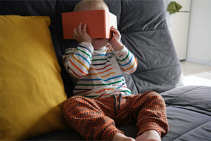A child in striped outfit playing with a cardboard VR set on a sofa, related to petty grudges theme.