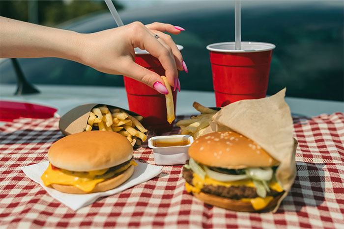 A hand with pink nails holds a fry over burgers and drinks on a picnic table, illustrating a lighthearted grudge display.