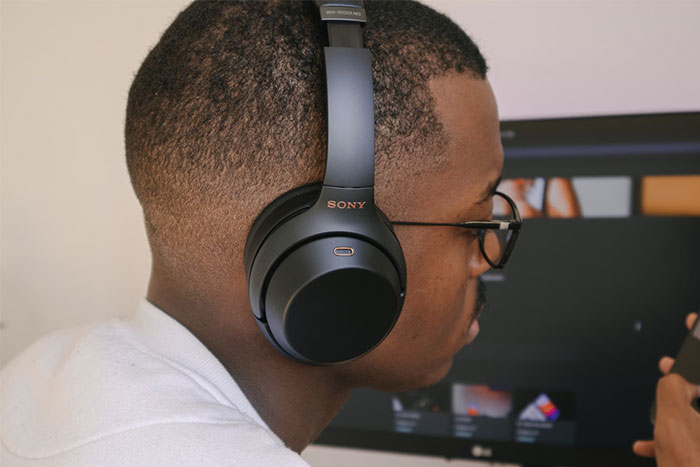 Man wearing Sony headphones, holding a phone, in front of a computer screen.