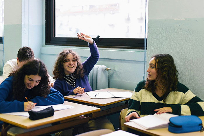 Students sitting at desks in a classroom, one raising a hand, possibly discussing petty grudges.