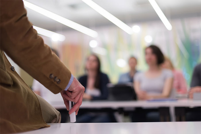 A lecturer in a brown jacket holds chalk, facing a blurred audience, symbolizing personal grudges in a classroom setting.