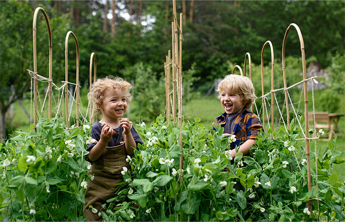 Two children playing joyfully in a green garden, surrounded by tall plants and trellises.