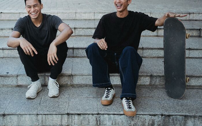 Two friends laughing on steps, capturing a moment of hilarious overheard conversations, with a skateboard beside them.