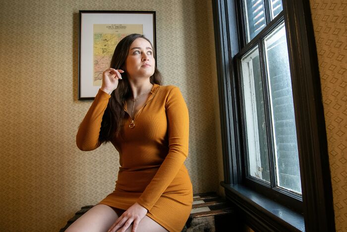 Person in a mustard dress sitting by a window, pondering rumors.