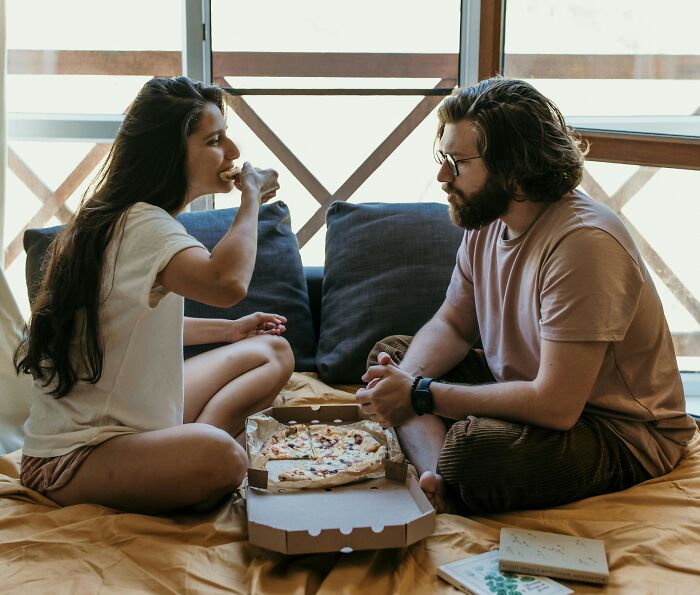 Couple sharing a pizza on a bed, surrounded by cushions, showcasing love and togetherness.