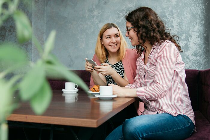 Two women in a café smiling and chatting, sharing a funny overheard conversation over coffee.