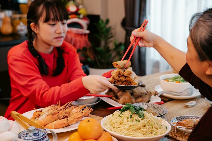 Two people sharing spring rolls with chopsticks, surrounded by a variety of dishes, highlighting cultural differences in dining.
