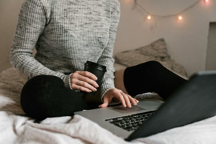 Person in cozy clothes sitting on bed with laptop and coffee cup, enjoying the perks of not having to go to the office