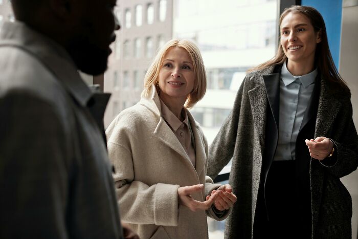 Two women in coats smiling and chatting with a man in an office setting, illustrating hilarious overheard conversations.