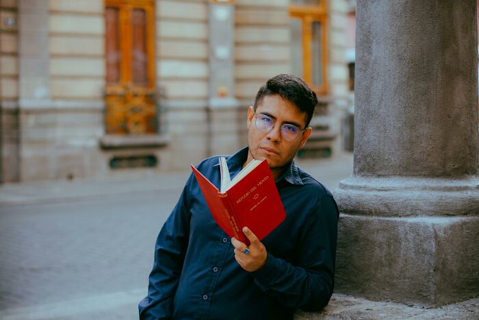 Man in blue shirt, wearing glasses, reading a red book by a column, illustrating genetic blessings and weaknesses themes.