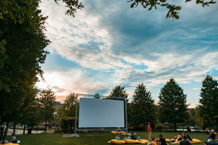 Outdoor screen setup in a park, surrounded by trees and people relaxing on the grass, for a weird government project event.