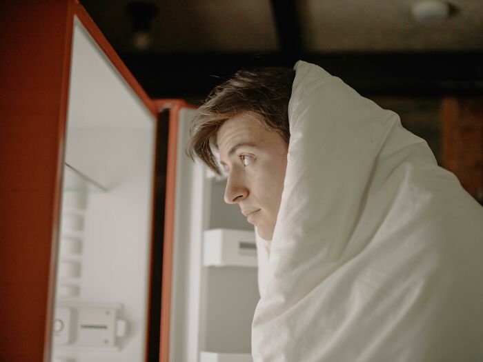 Young man wrapped in a blanket looking into a fridge, illustrating perks of not having to go to the office at home.