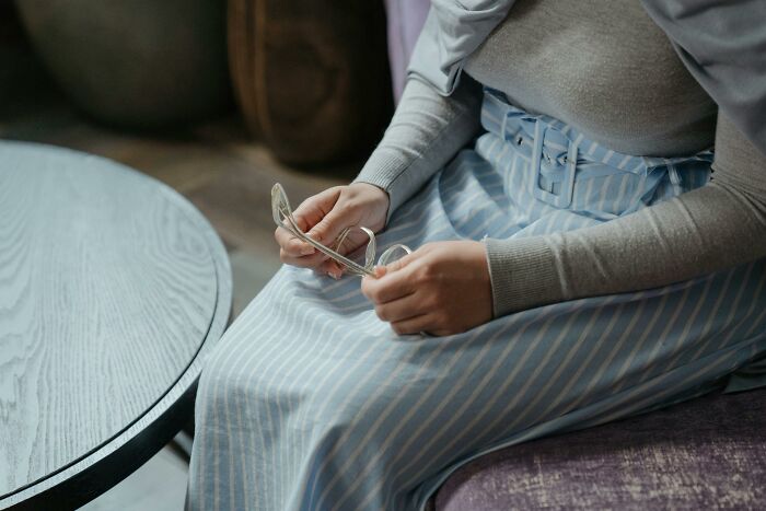 Person holding glasses, seated in a striped blue skirt, possibly recalling hilarious conversations.