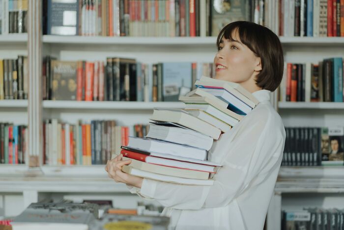 Person in a library holding a large stack of books, surrounded by shelves filled with more books.