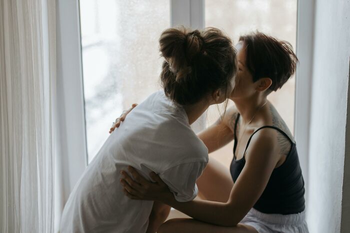 Women sharing an intimate moment by a window, demonstrating connection and affection.