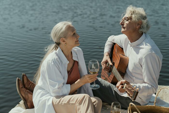 Senior couple enjoying eye-opening moments by a lake, with wine and a guitar.