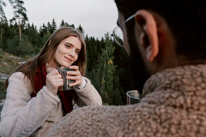 Woman having an eye-opening moment, smiling with a metal cup, wearing a fleece jacket in an outdoor forest setting.