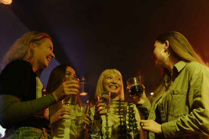 Four women laughing and holding drinks at a party, possibly sharing hilarious overheard conversations.