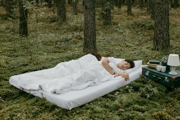 Person sleeping on a mattress in a forest with a table and lamp nearby, showcasing brilliant life hacks.