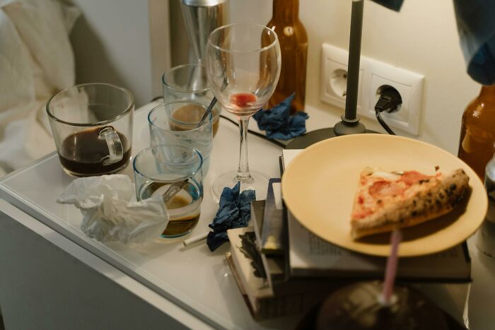 Messy bedside table with leftover pizza, empty glasses, and clutter, reflecting weird personal habits.