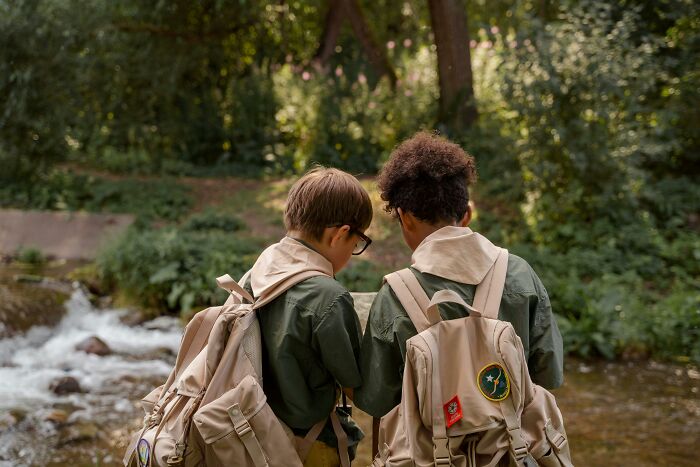 Two scouts by a stream sharing hilarious overheard conversations, wearing green shirts and backpacks in a forest setting.