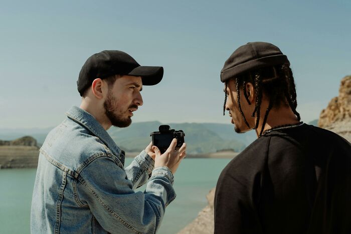 Two men outdoors by a lake, one holding a camera, discussing life cheat codes for a smarter and more enjoyable experience.