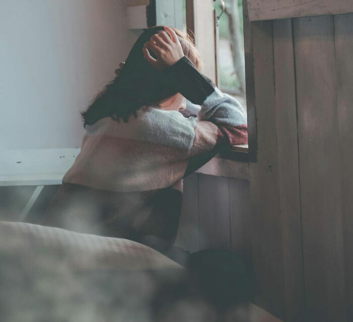 Person in a sweater leans on a windowsill, looking outside, pondering a creepy family memory.