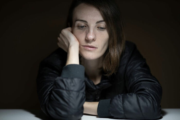 Person with a prominent nose, resting their chin on their hand, wearing a dark jacket in a dimly lit room.