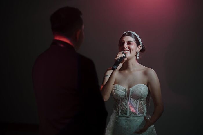 Bride singing at wedding reception with microphone, wearing a lace gown; facing a man in a dark suit.