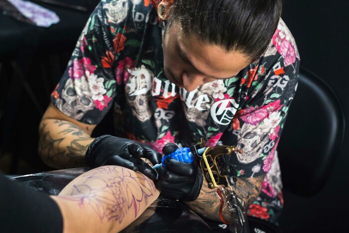 Tattoo artist in floral shirt working on an arm design in a studio setting.