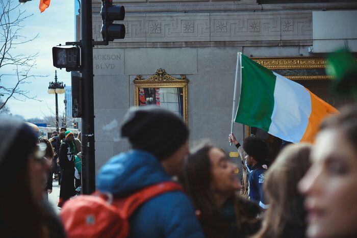 A person holds an Irish flag during a parade, with people conversing humorously in the foreground.